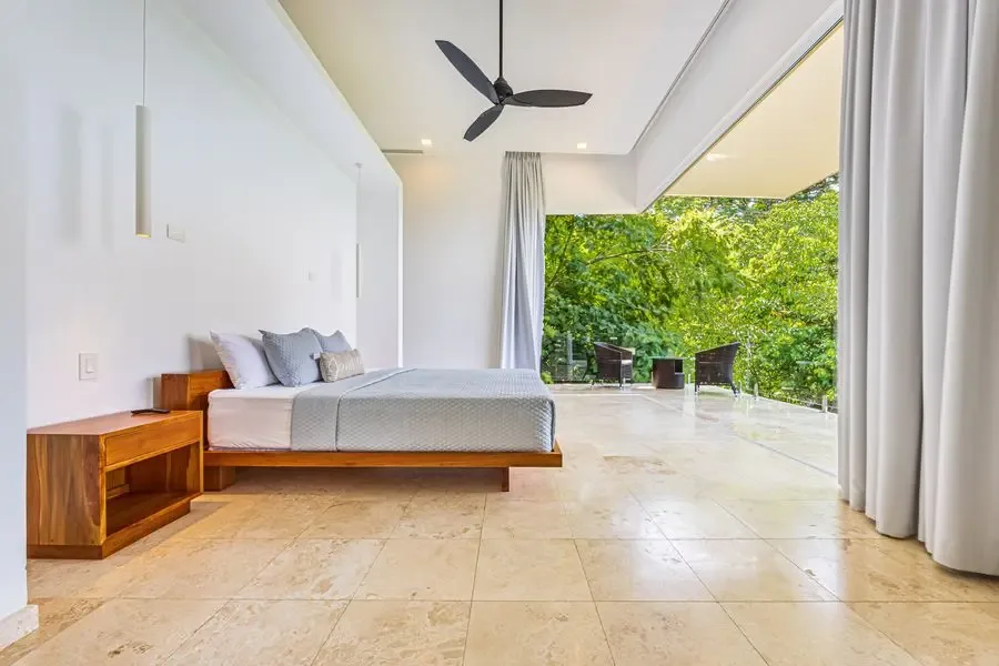 A bright bedroom opens onto a leafy terrace, with a bed and ceiling fan.