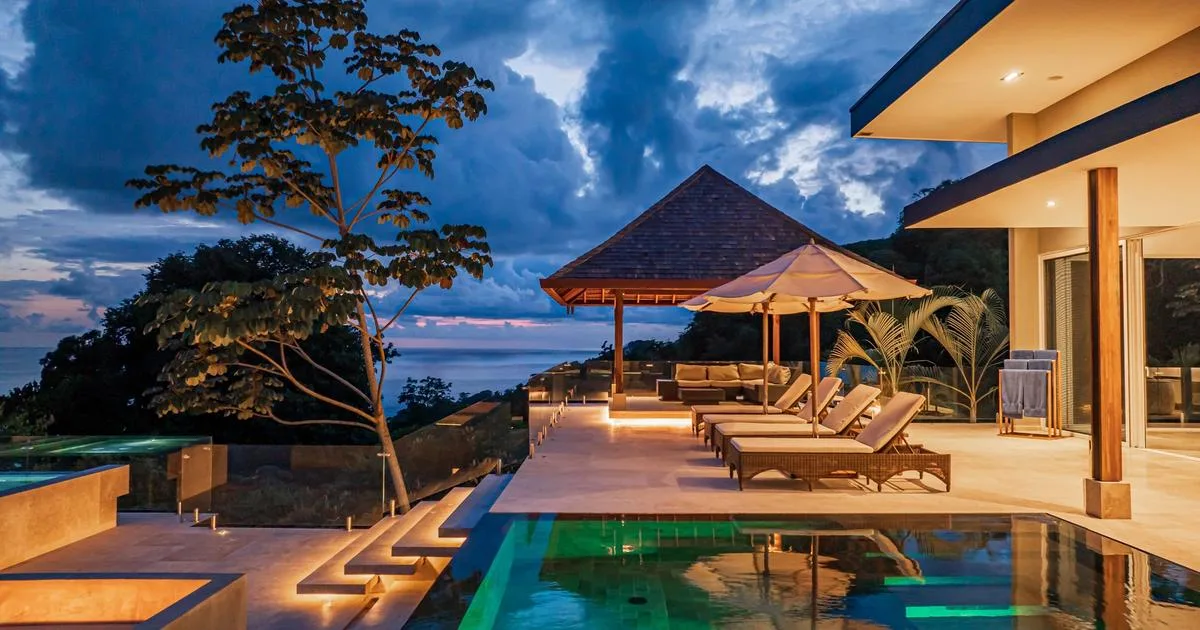 An outdoor pool beside a modern house, with lounge seating under umbrellas.