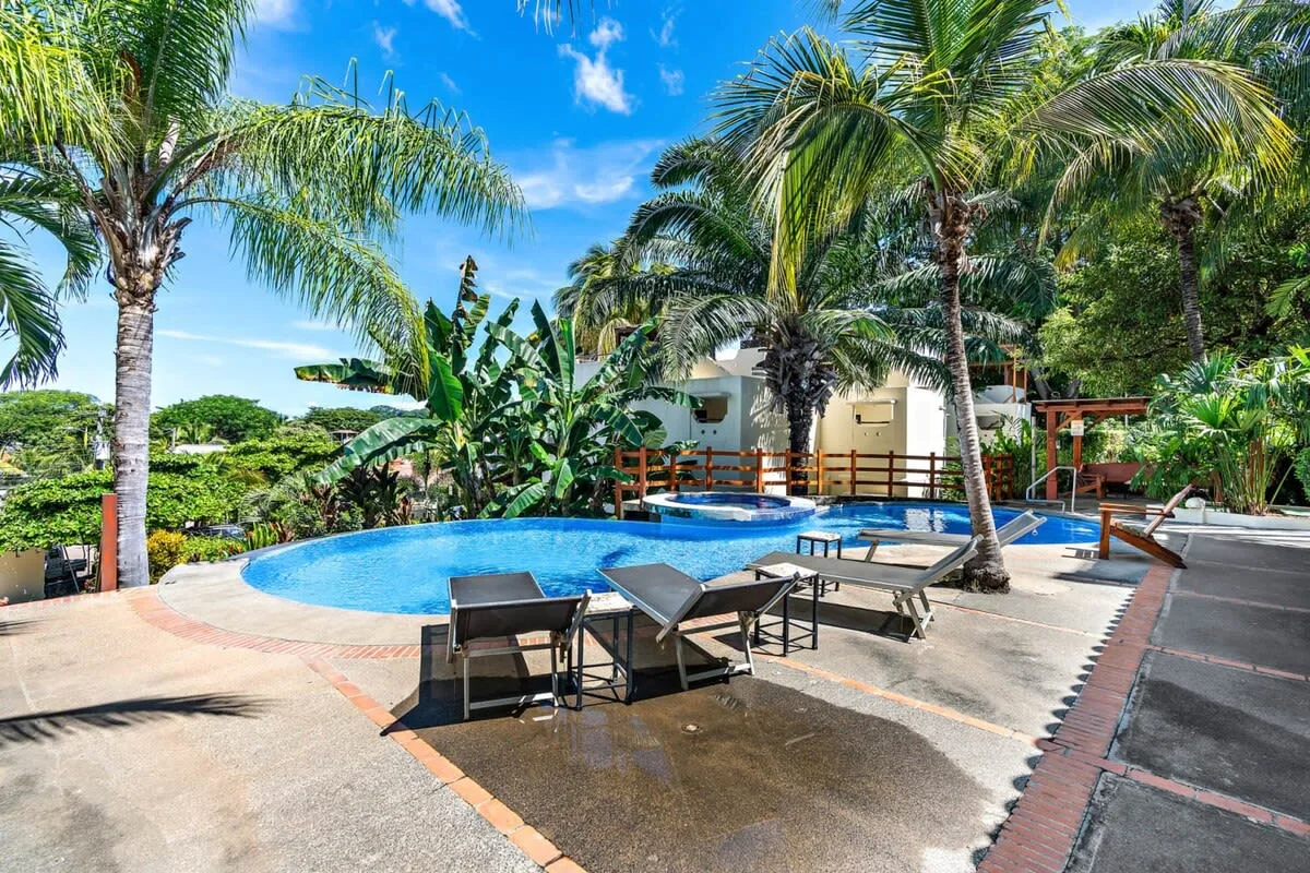 Outdoor swimming pool surrounded by palm trees, with lounge seating along the deck.
