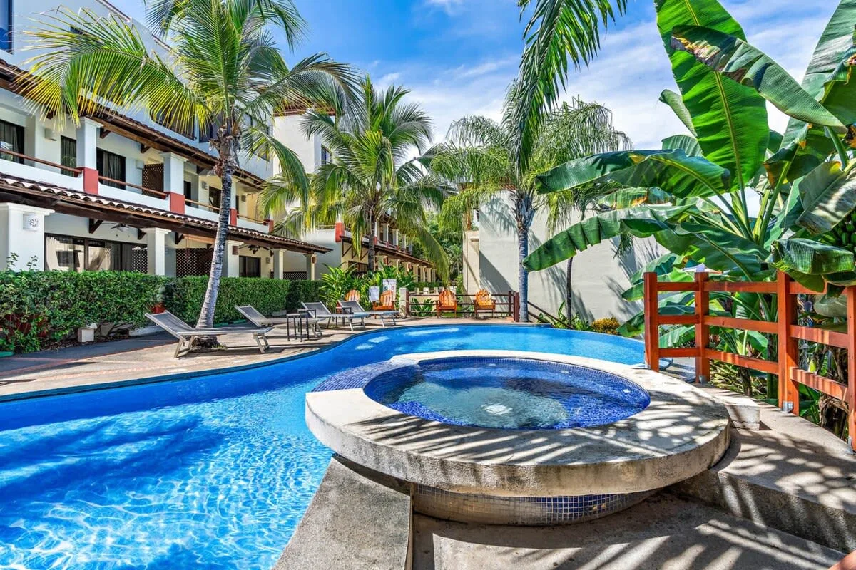 A tropical outdoor pool and round spa are surrounded by palm trees and lounge seating.