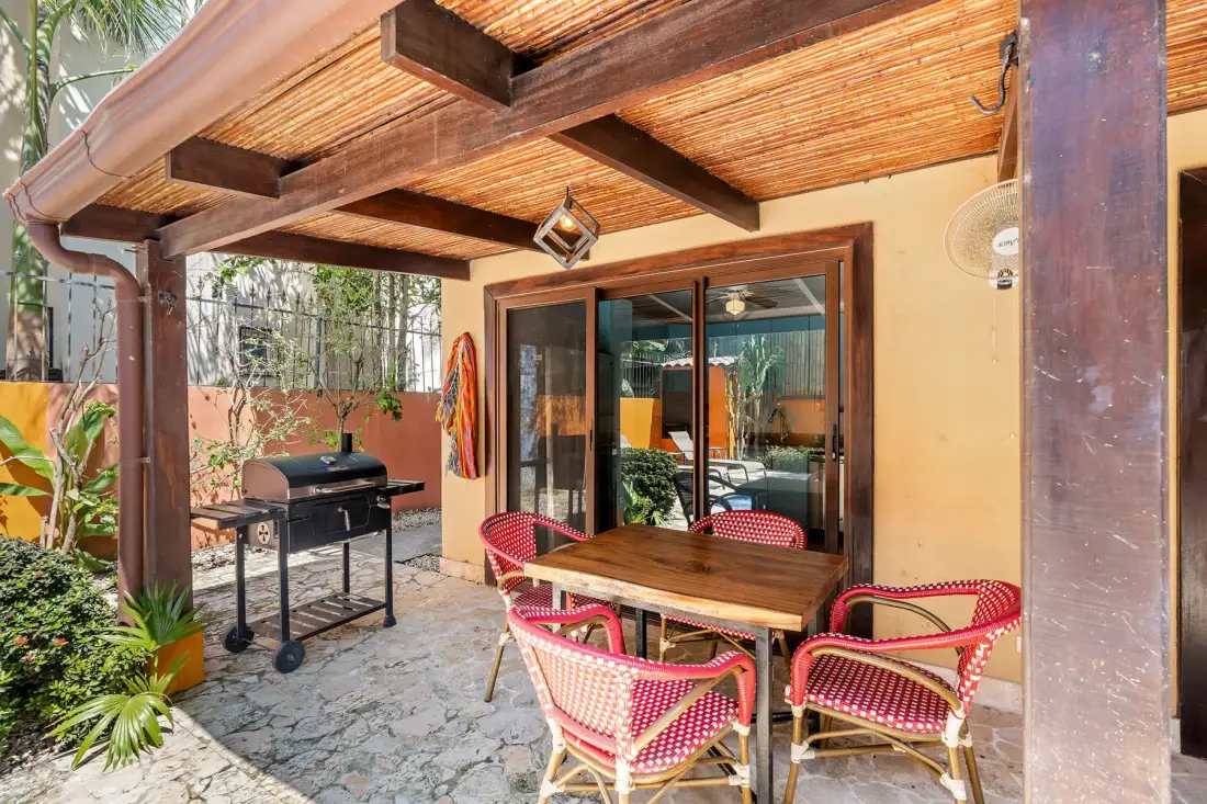 A covered patio has a wooden dining table, woven seating, and a grill.