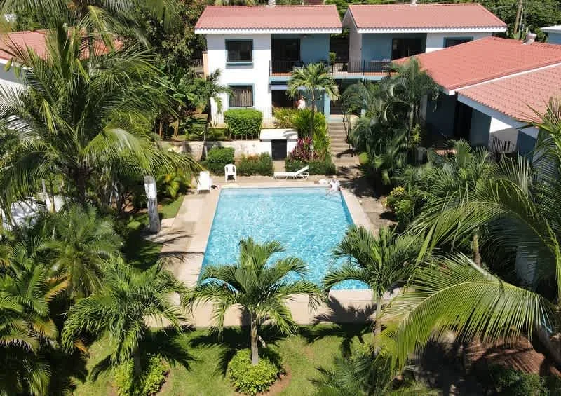 A swimming pool sits among tropical greenery beside low-rise buildings.