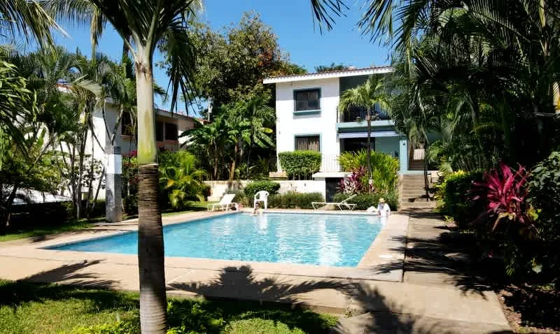 A swimming pool sits among palm trees in front of a white building.