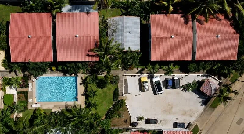 An aerial view of red-roofed buildings, a swimming pool, and a parking area among palm trees.