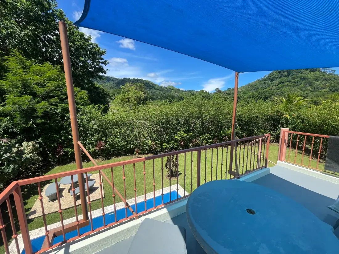 A shaded outdoor patio overlooks green hills, with a round table beside the railing.