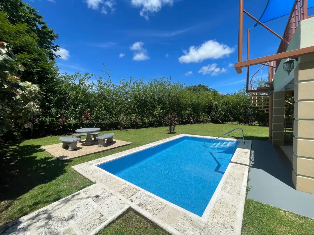 A backyard swimming pool is surrounded by grass, shrubs, and a small patio table.