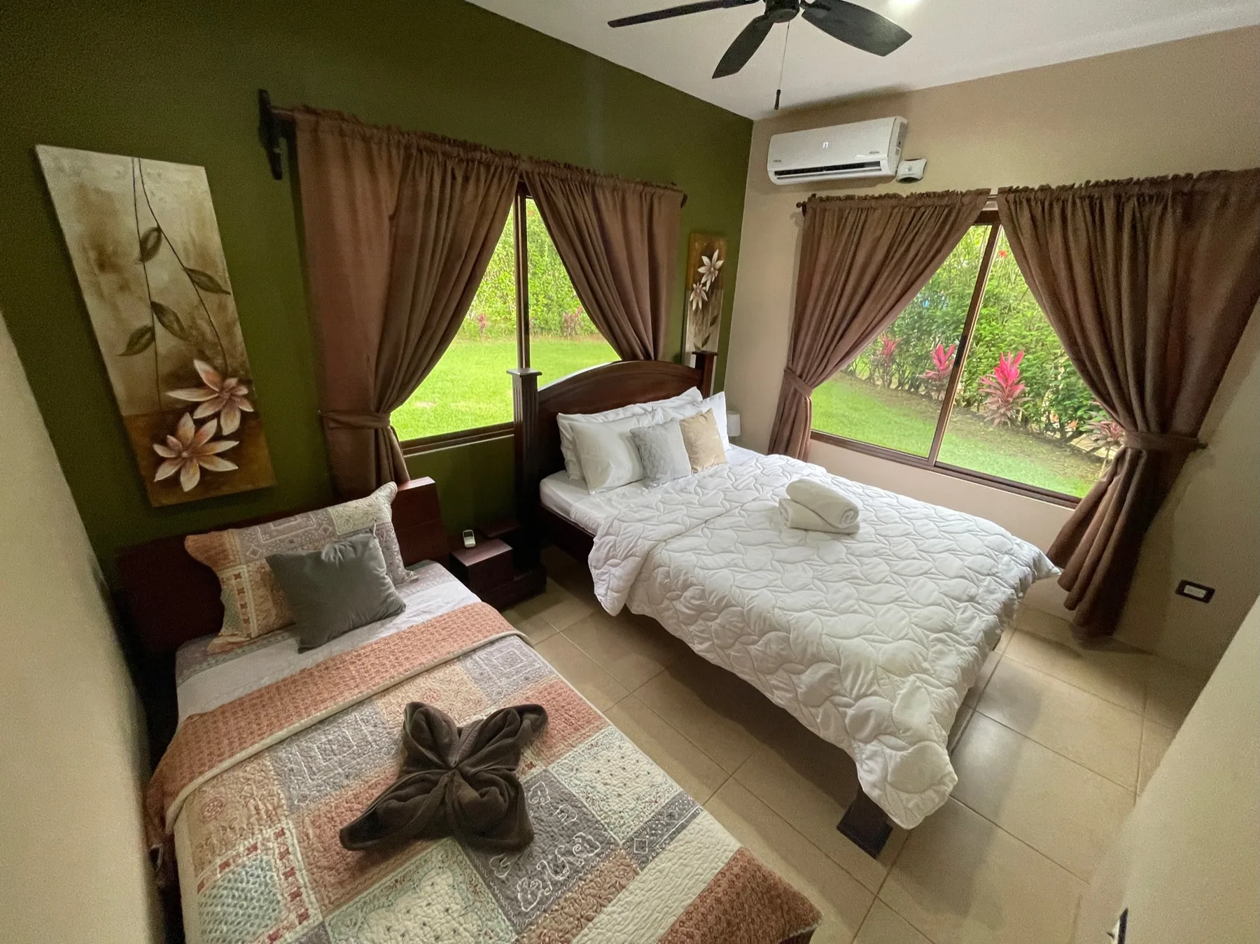 A bedroom with two beds, brown curtains, and windows overlooking greenery.