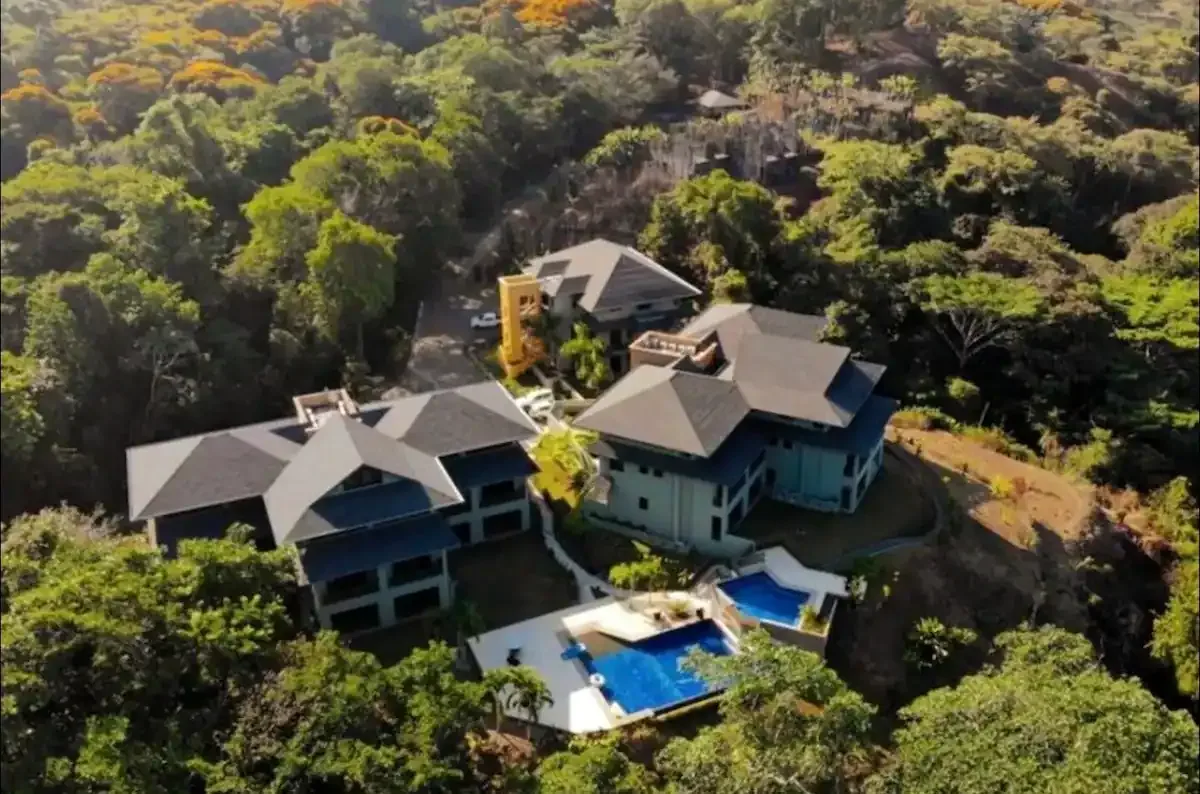 An aerial view of several buildings surrounded by dense greenery, with a swimming pool.