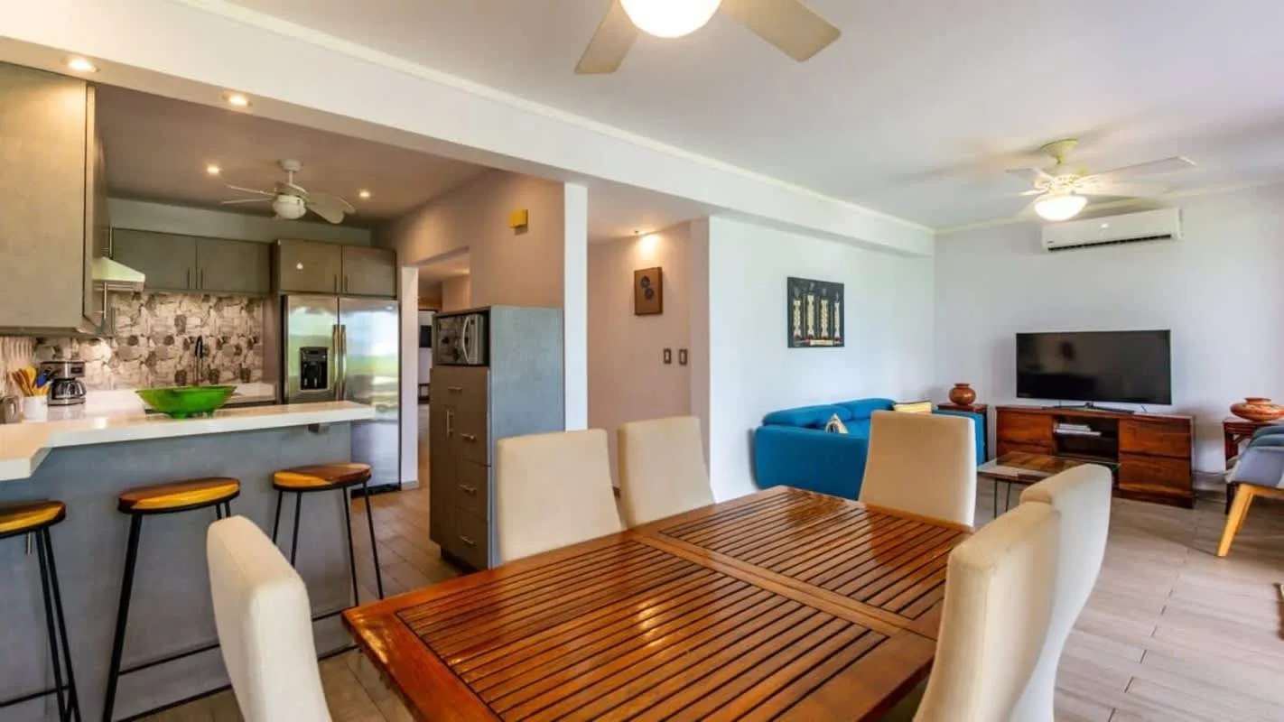 A dining and living area with a wooden table, kitchen counter, and blue seating.