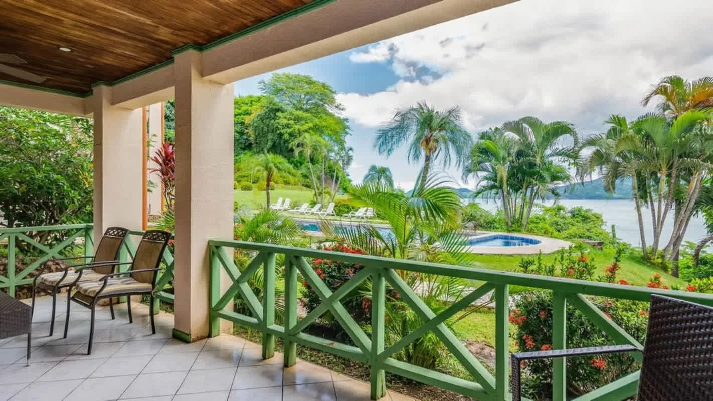 A covered patio overlooks a swimming pool, tropical greenery, and the water.