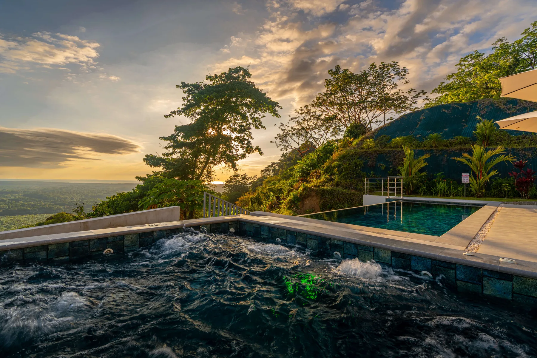 Una piscina al aire libre mira hacia una ladera arbolada bajo un cielo de colores al atardecer.
