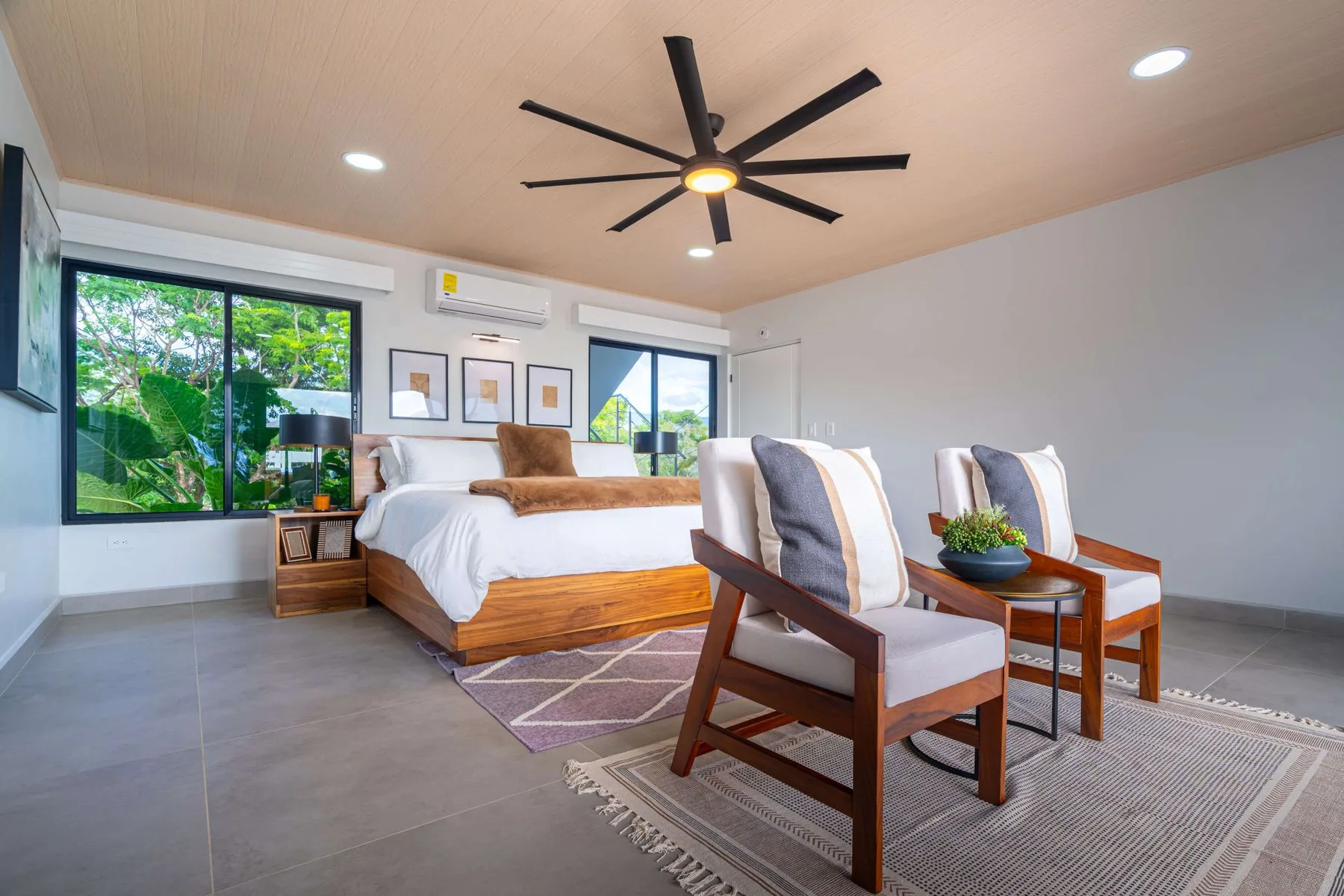 Un dormitorio luminoso con una cama y dos sillones junto a las ventanas.