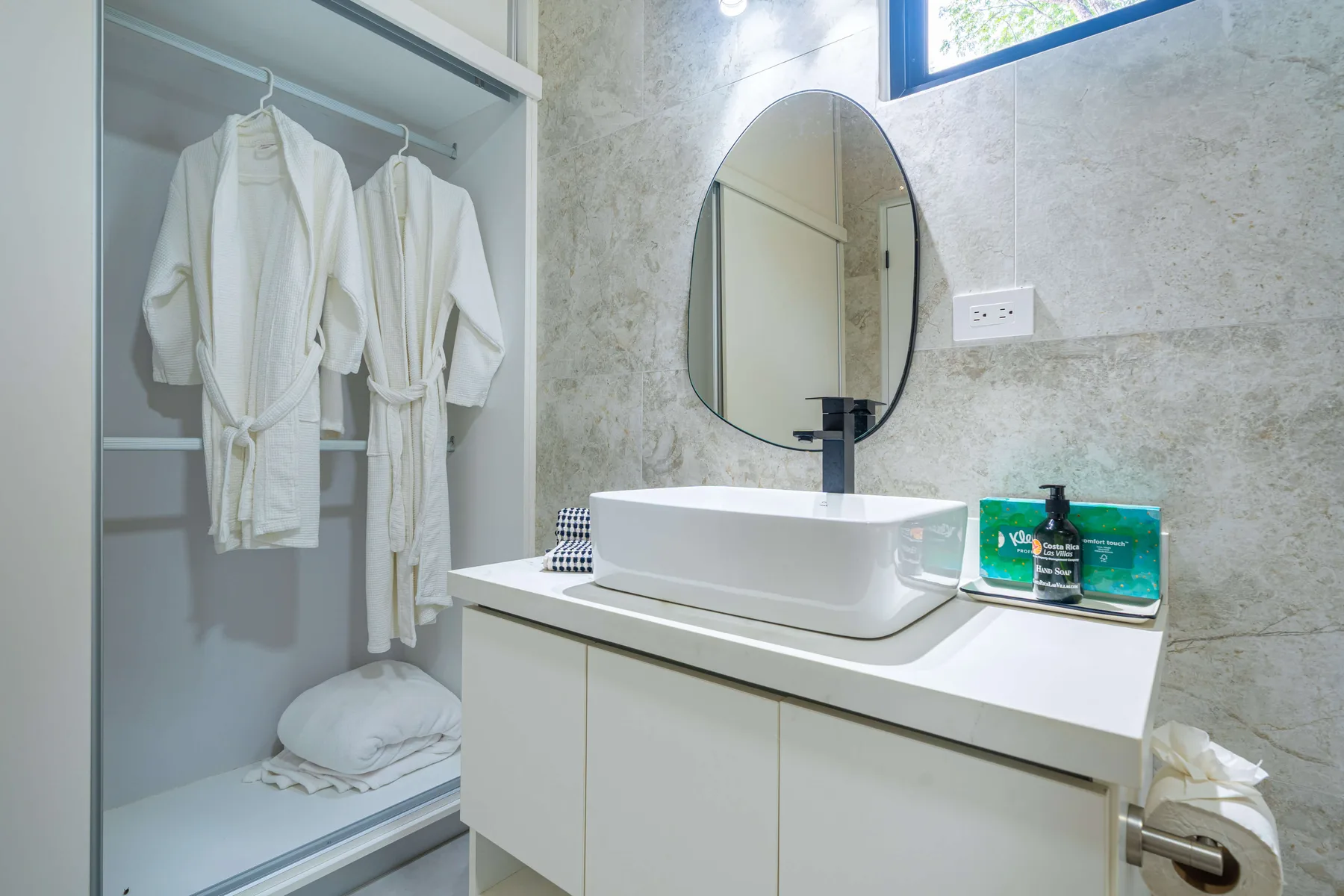 Un lavabo de baño con un lavamanos sobrepuesto, espejo ovalado y espacio abierto para albornoces.