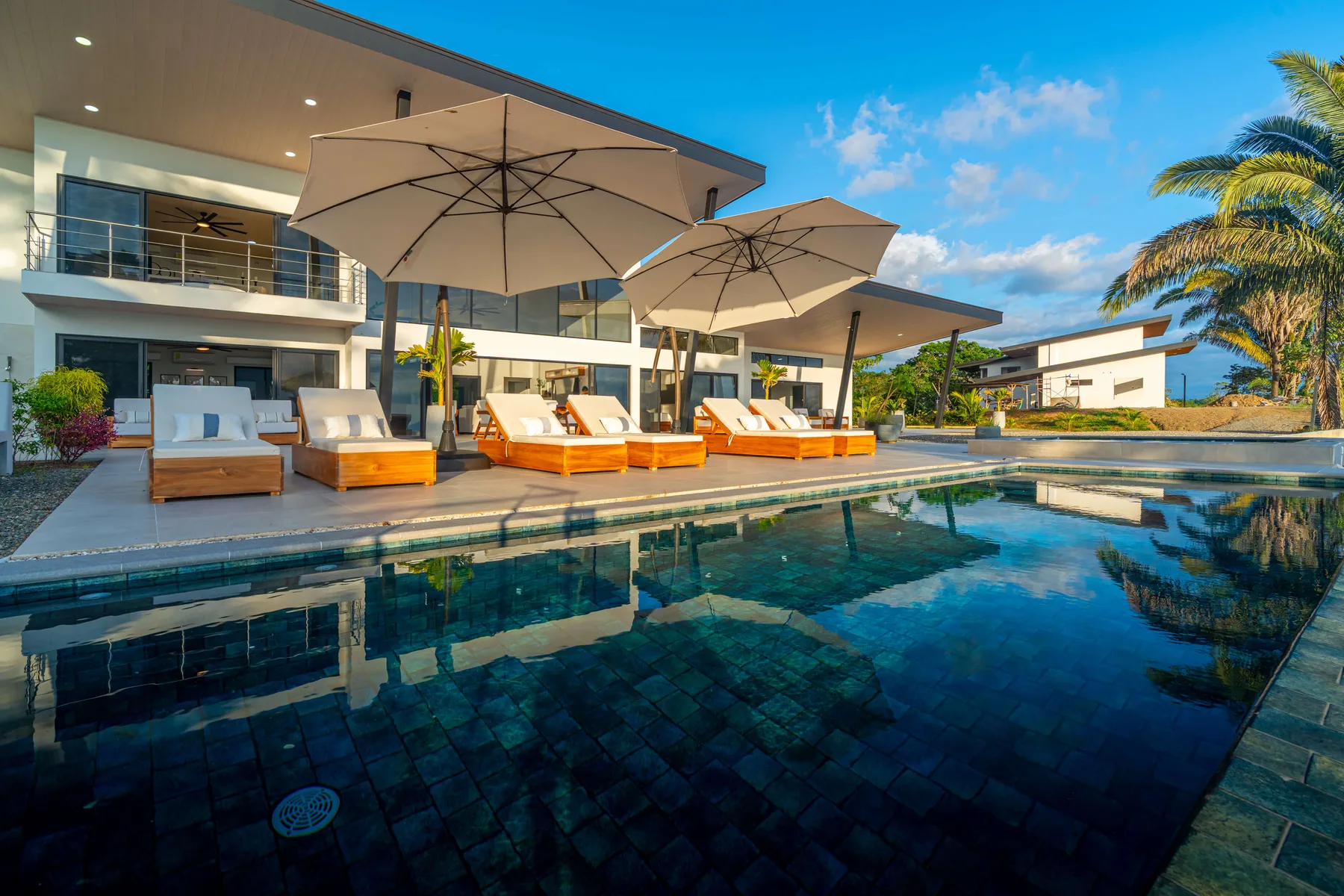 Una piscina junto a una casa moderna, con asientos de descanso y sombrillas.