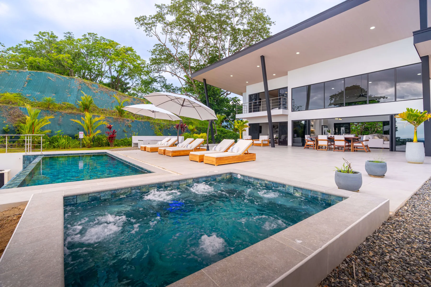 Una piscina exterior y un jacuzzi con burbujas están junto a una casa moderna con patio cubierto.