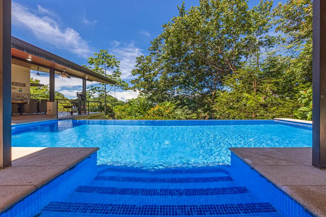 An outdoor swimming pool beside a glass-walled pavilion and leafy trees.