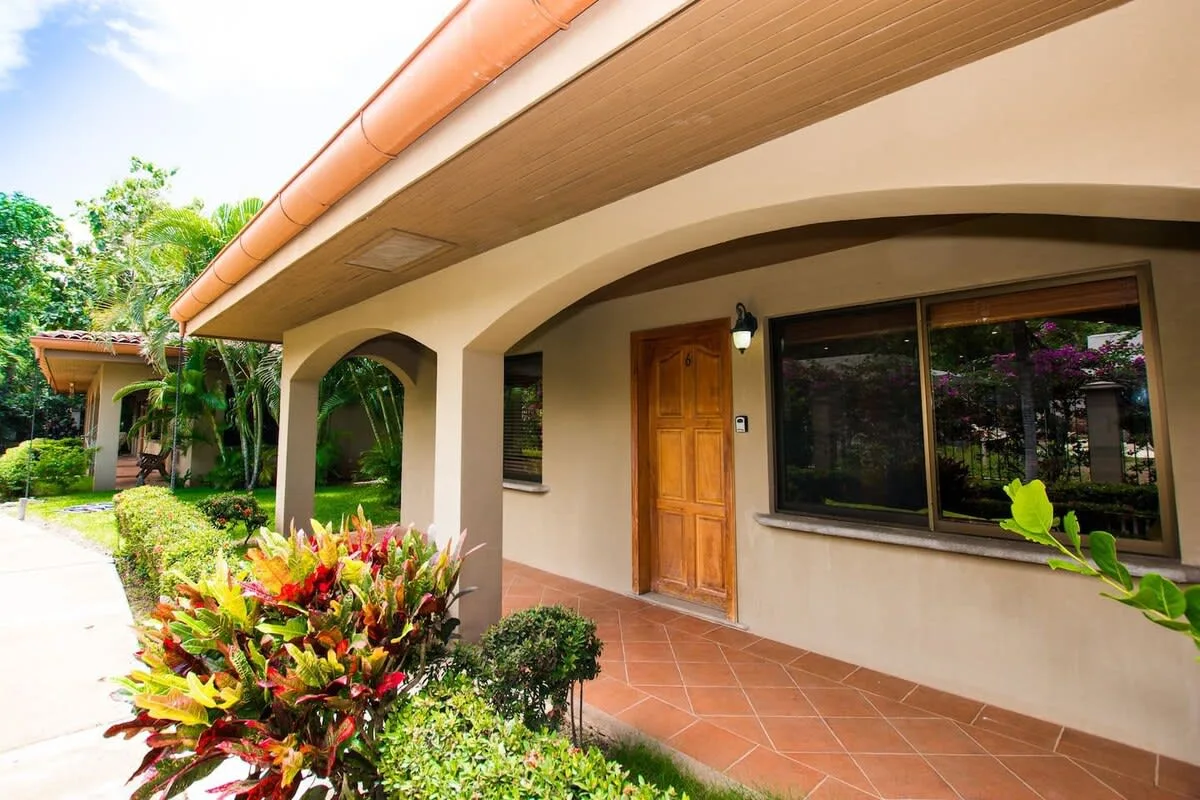 A covered exterior walkway runs beside a building, with a wooden door and garden plants.