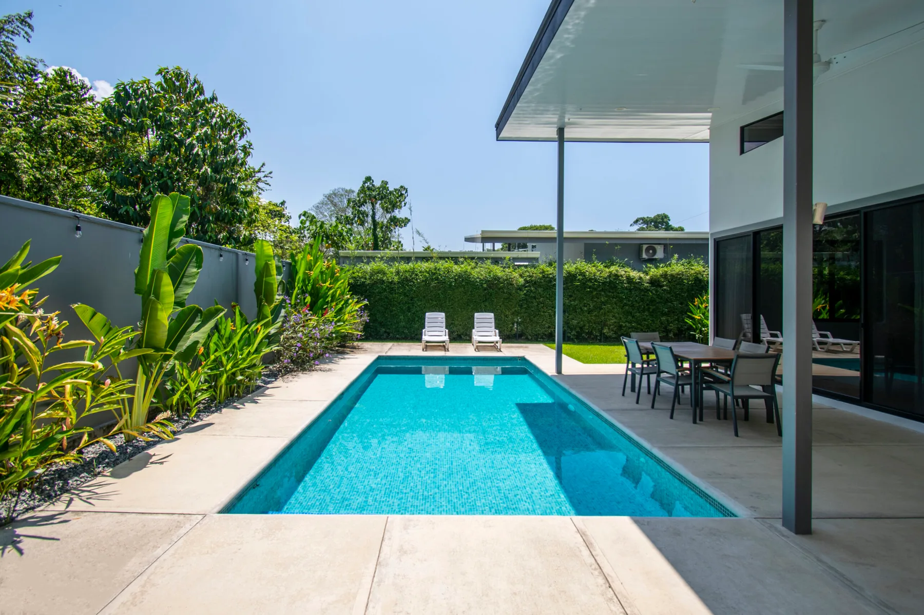 A rectangular swimming pool sits beside a modern patio with outdoor seating and greenery.