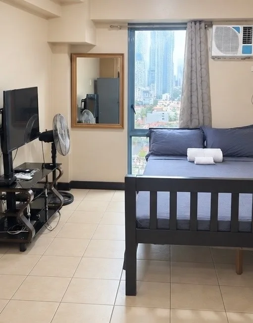 Un dormitorio con una cama oscura junto a una ventana amplia con vistas a edificios de la ciudad.