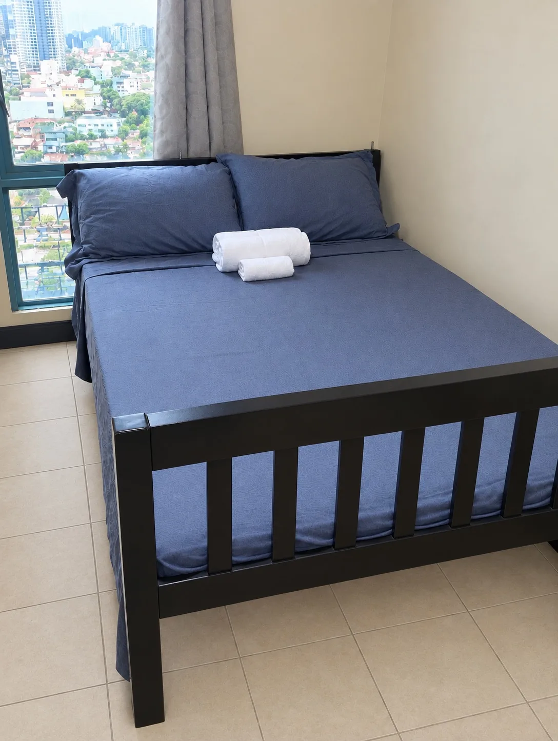 Un dormitorio con una cama oscura y toallas blancas dobladas junto a una ventana.