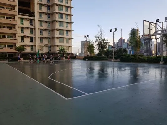 Una cancha de baloncesto al aire libre junto a un edificio alto, con edificios urbanos al fondo.