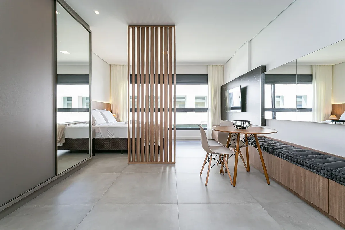 A bright bedroom and seating area are divided by a vertical wooden slat screen.