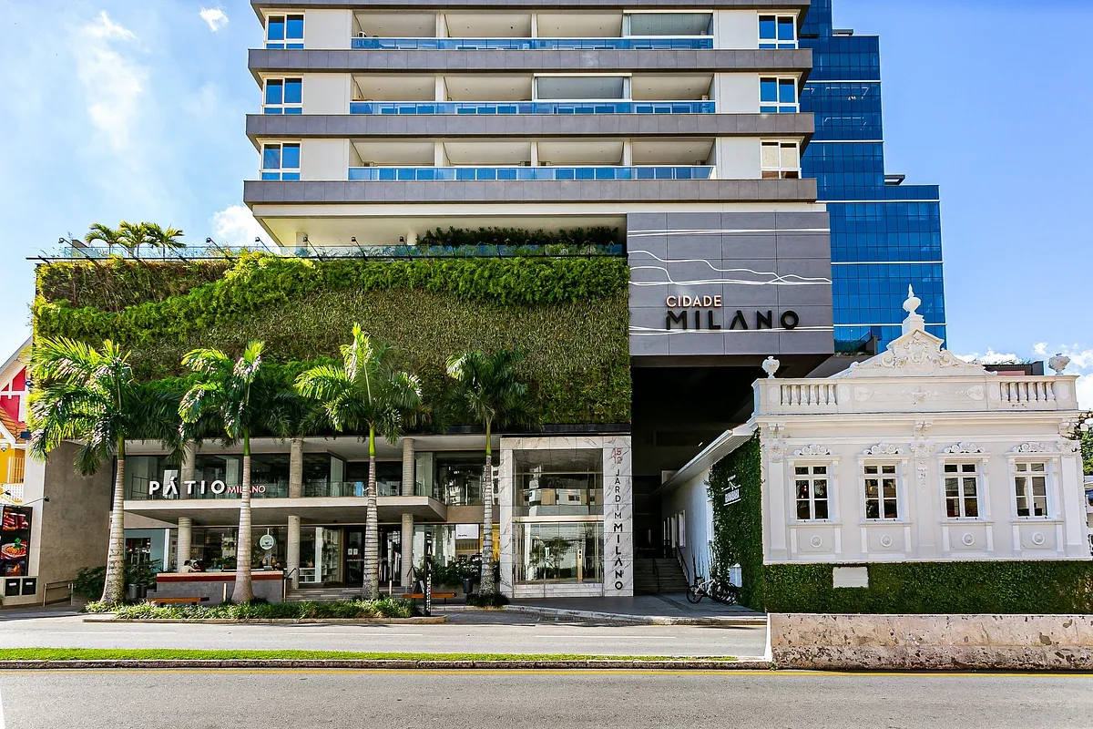 A modern multi-story building with balconies rises behind a leafy storefront.