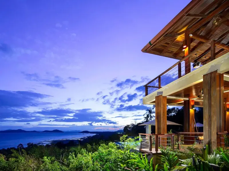 A modern villa overlooks the sea at dusk, with a lit balcony and surrounding greenery.
