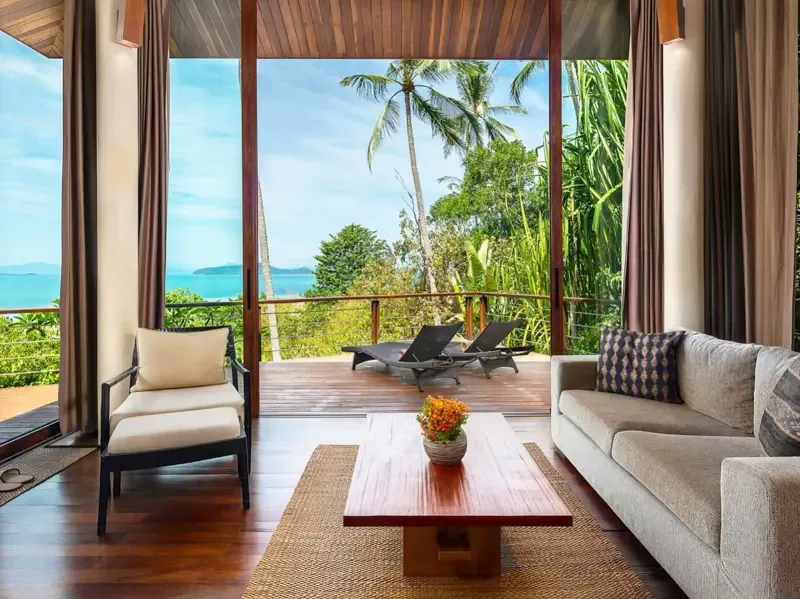 A bright living room opens onto a deck with lounge seating and a tropical view.
