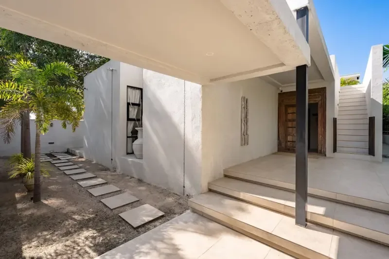 A white modern house entrance opens onto a paved walkway beside a small garden.