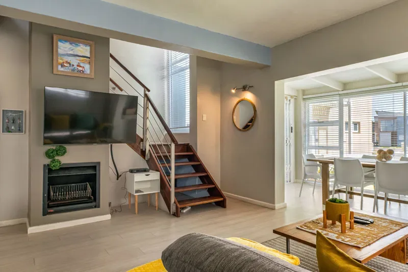 Una sala de estar con televisor, chimenea y escalera junto a un comedor luminoso.