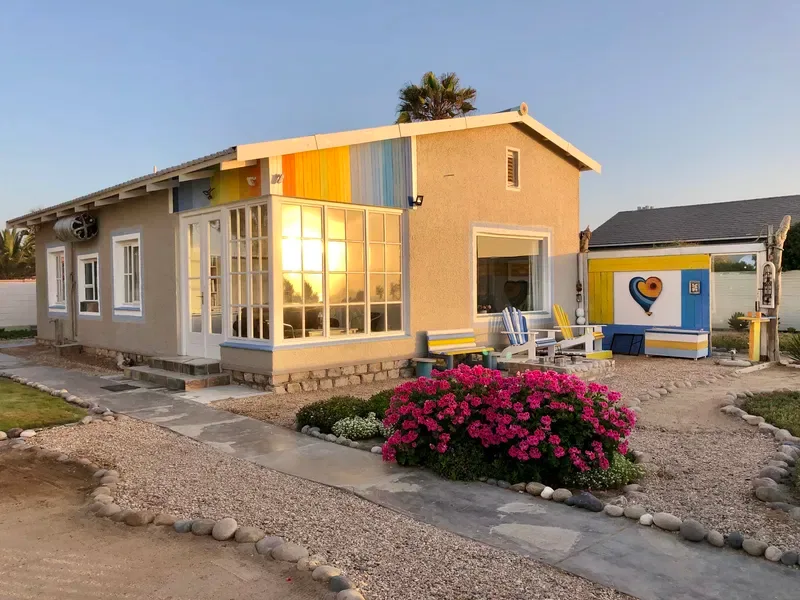 Una casa pequeña con un porche cerrado iluminado por el sol y arbustos en flor al frente.