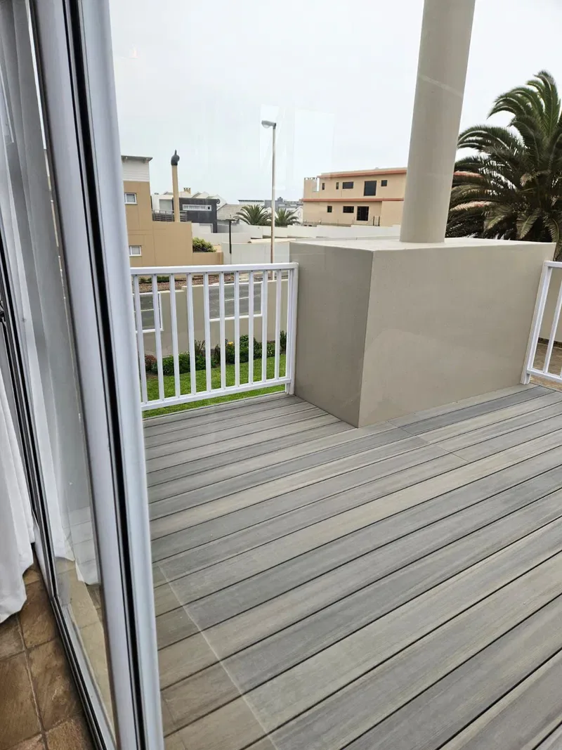 A balcony with gray decking and white railings overlooks nearby buildings and palm trees.