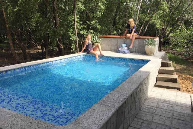 Una piscina al aire libre rodeada de árboles, con personas sentadas junto al agua.