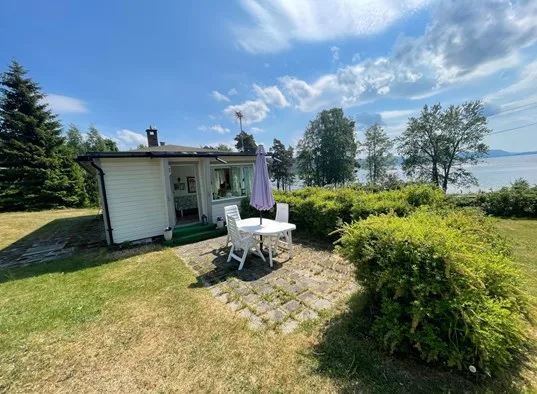 A small white cottage with outdoor seating stands beside a garden overlooking the water.