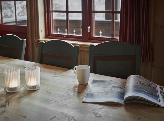 A dining table with a mug and open book sits beside a row of windows.