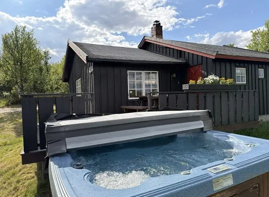 Una cabaña de madera oscura se encuentra detrás de una bañera de hidromasaje al aire libre.