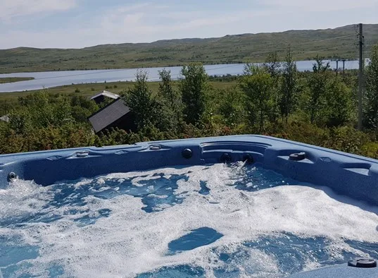 Una bañera de hidromasaje al aire libre tiene vistas a árboles, un lago y colinas.