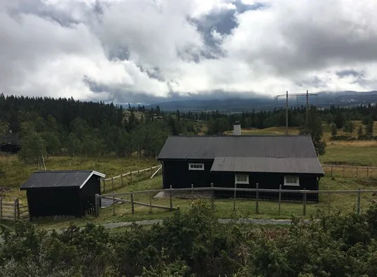 A dark cabin and small shed sit in a fenced rural landscape beneath cloudy skies.