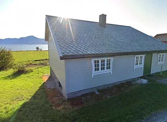 A gray cottage stands beside a grassy lawn, with water and hills in the background.