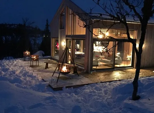 A glass-walled cabin glows warmly in a snowy outdoor setting at dusk.