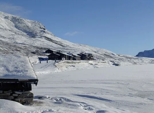 Snow-covered mountain cabins stand beside a broad frozen, snow-covered landscape.