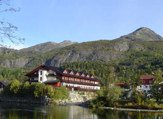 Un alojamiento de montaña se alza junto a un lago tranquilo, con laderas boscosas al fondo.