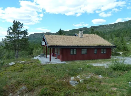 A red cabin stands in an open mountain landscape beneath a partly cloudy sky.