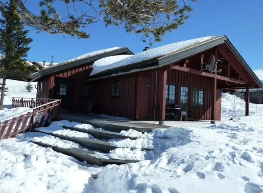 A dark wooden cabin stands in a snowy landscape beneath a clear blue sky.