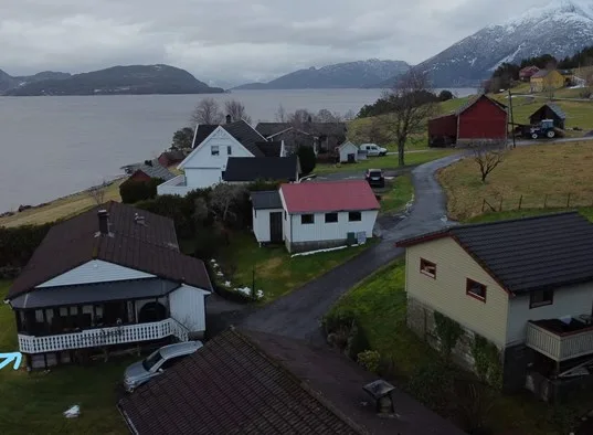 Houses line a curving road beside a broad lake, with mountains in the distance.