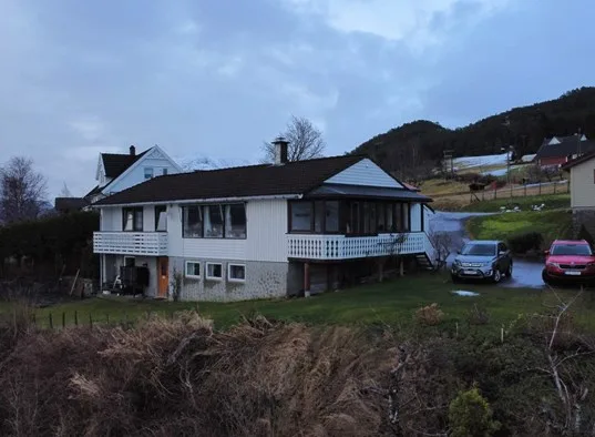 A white house with a dark roof sits beside a grassy hillside.