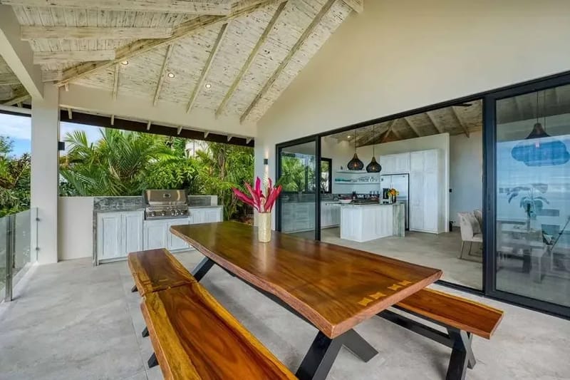 Cocina abierta y una larga mesa de comedor con bordes naturales bajo un techo abovedado de bambú