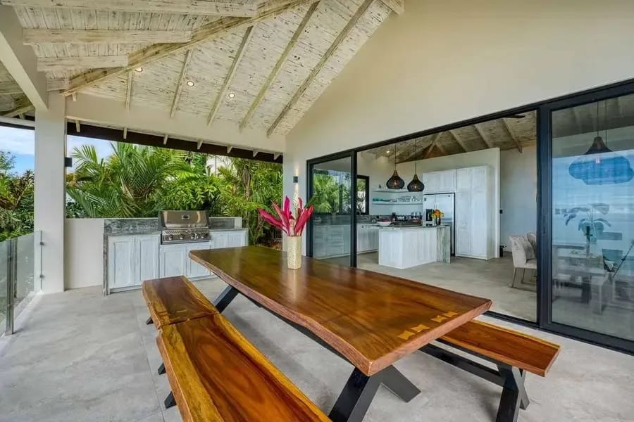 Cocina abierta y una larga mesa de comedor con bordes naturales bajo un techo abovedado de bambú