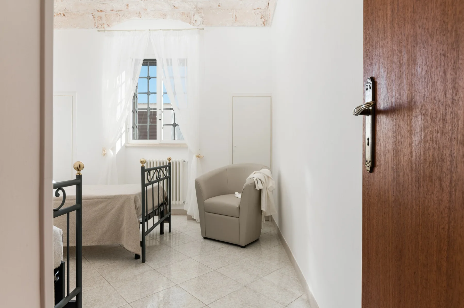 A bright bedroom with a bed, an armchair, and a window with sheer curtains.